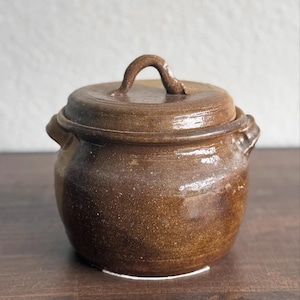 May include: A small, brown ceramic pot with a lid. The pot has a rounded body, two small handles, and a curved handle on the lid. The ceramic has a speckled appearance and a glossy finish. The pot is sitting on a wooden surface.