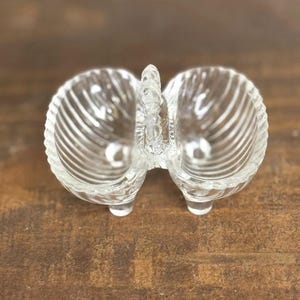 May include: Clear glass double dish with a seashell design. The dish has two compartments and a handle in the center. The dish sits on three small feet. The glass has a ribbed texture.