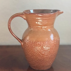 May include: A ceramic pitcher with a textured lower half and a smooth, glossy upper half. The pitcher is a warm, earthy orange color with a curved handle and a spout for pouring. The pitcher is likely used for serving beverages.