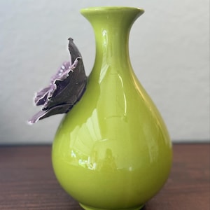 Puede incluir: Un jarr&oacute;n de cer&aacute;mica verde lima brillante. Una flor tridimensional de color p&uacute;rpura oscuro est&aacute; adherida al lateral. El jarr&oacute;n tiene un cuerpo redondeado y un cuello delgado, ideal para flores.