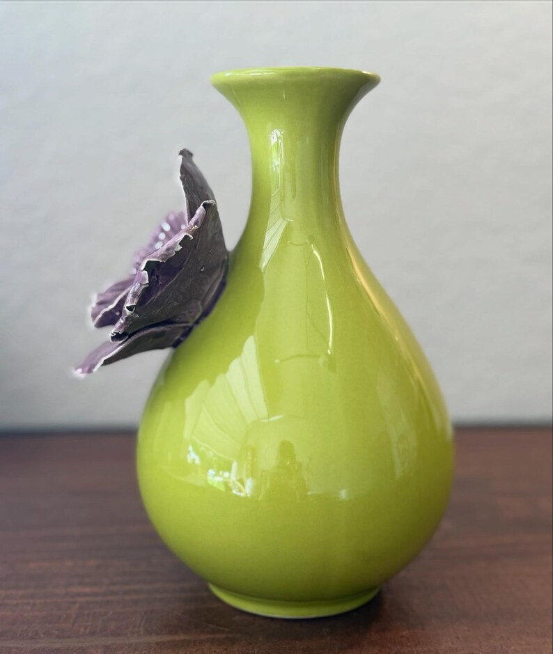 Puede incluir: Un jarr&oacute;n de cer&aacute;mica verde lima brillante. Una flor tridimensional de color p&uacute;rpura oscuro est&aacute; adherida al lateral. El jarr&oacute;n tiene un cuerpo redondeado y un cuello delgado, ideal para flores.
