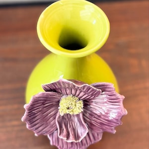 Puede incluir: Un jarr&oacute;n de cer&aacute;mica verde lima con un cuerpo bulboso y una abertura acampanada. Una gran flor morada texturizada con un centro amarillo est&aacute; adherida al frente, agregando un elemento decorativo. El jarr&oacute;n es probablemente para arreglos florales.