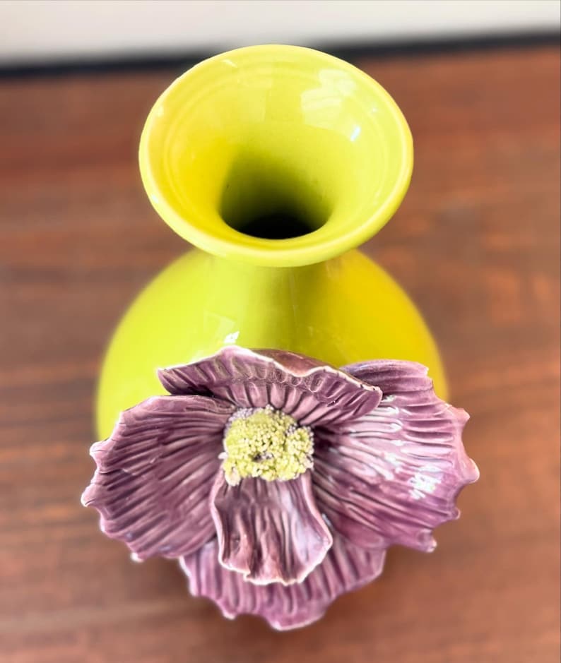 Puede incluir: Un jarr&oacute;n de cer&aacute;mica verde lima con un cuerpo bulboso y una abertura acampanada. Una gran flor morada texturizada con un centro amarillo est&aacute; adherida al frente, agregando un elemento decorativo. El jarr&oacute;n es probablemente para arreglos florales.