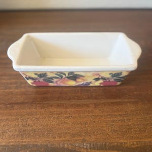 May include: A small, rectangular ceramic dish with a white interior and a decorative exterior. The exterior features a yellow background with a pattern of red and purple fruits and green leaves. The dish has small handles on each end.