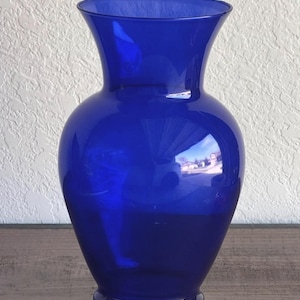 May include: A vibrant cobalt blue glass vase with a wide, rounded body and a flared top. The vase reflects the surrounding environment, showcasing a glossy finish. This decorative vase is suitable for floral arrangements or as a standalone accent piece.