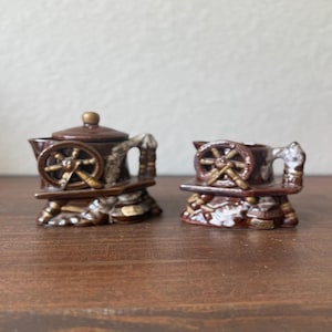May include: A pair of brown ceramic decorative pieces, one with a lid, resembling miniature water wheels. The pieces have a rustic design with a wheel and arch details, set on a textured base. The color palette includes shades of brown and beige.