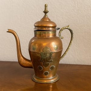 May include: Copper teapot with a long spout and ornate handle. The body features brass accents and decorative floral patterns. The lid is topped with a decorative finial. A vintage kitchenware item.
