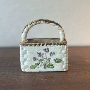 May include: A ceramic basket-shaped planter with a woven texture and a curved handle. The planter is painted white with a floral design of purple flowers and green leaves. The rim and handle are accented with brown.