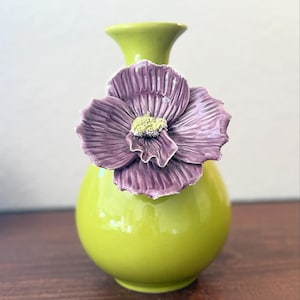 Puede incluir: Un jarr&oacute;n de cer&aacute;mica verde lima vibrante con un gran acento floral morado texturizado. La flor tiene un centro amarillo. El jarr&oacute;n tiene un cuerpo redondeado y un cuello delgado, creando una pieza decorativa llamativa.