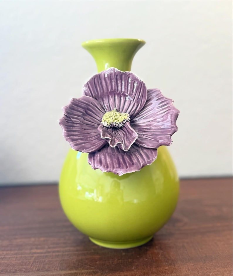 Puede incluir: Un jarr&oacute;n de cer&aacute;mica verde lima vibrante con un gran acento floral morado texturizado. La flor tiene un centro amarillo. El jarr&oacute;n tiene un cuerpo redondeado y un cuello delgado, creando una pieza decorativa llamativa.