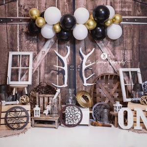 May include: A rustic-themed photo backdrop with a wooden barn door design. The scene includes white, black, and gold balloons, decorative antlers, and various props like a window frame, lanterns, and the word "ONE" in large letters. The backdrop is ideal for a birthday celebration.