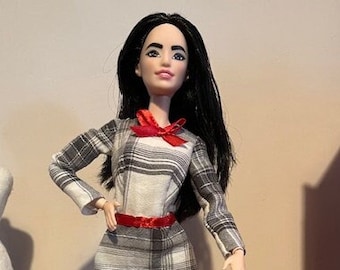 May include: A fashion doll with long black hair wearing a plaid dress with a red bow and red shoes. The doll is standing on a clear plastic doll stand.