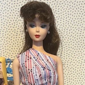 Può includere: Una bambola con capelli castani raccolti in una coda di cavallo alta, che indossa un abito monospalla con un motivo a righe rosse, bianche e blu con accenti di cuciture nere. L'abito è stretto in vita da una sottile cintura a nastro nero.