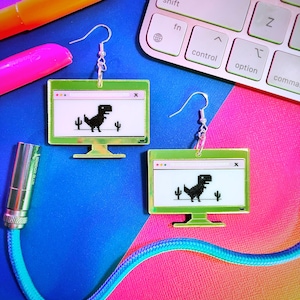 May include: A pair of gold-coloured earrings shaped like a computer screen with a pixelated dinosaur running on the screen. The earrings are on a pink and blue background.