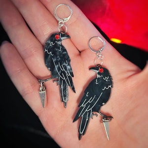 May include: A pair of black crow earrings with red eyes and silver accents. Each crow is perched on a silver dagger.