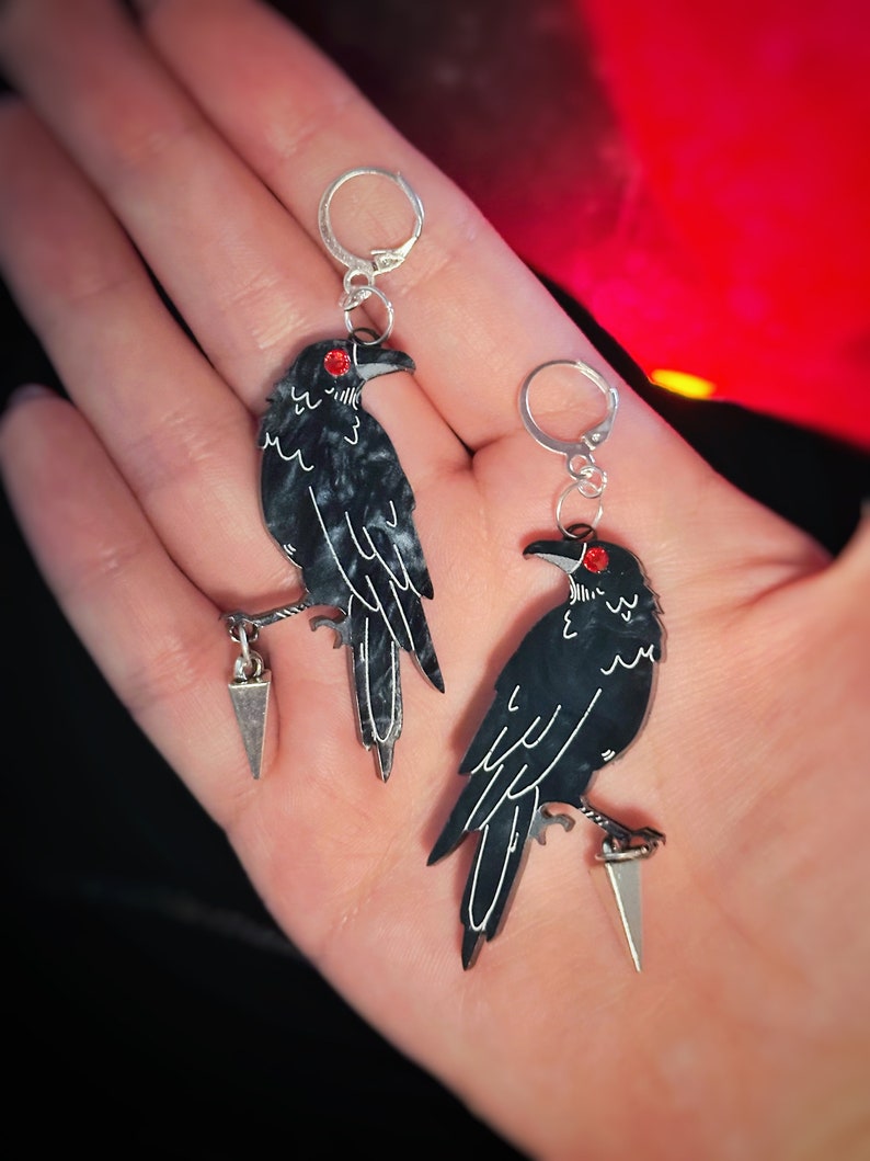 May include: A pair of black crow earrings with red eyes and silver accents. Each crow is perched on a silver dagger.