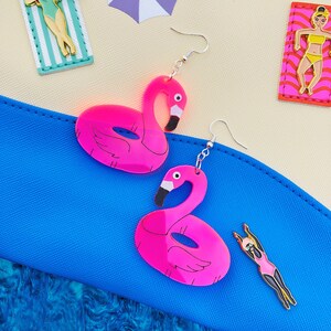 May include: A pair of pink flamingo pool float earrings. The earrings are made of acrylic and have a shiny finish. The earrings are hanging from silver hooks.