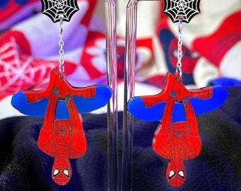May include: A pair of dangle earrings featuring a red and blue Spiderman figure hanging upside down from a black web charm. The earrings are displayed on a clear acrylic stand.