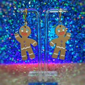 May include: A pair of gingerbread man earrings with white icing details and a red smile. The earrings are hanging from gold hooks on a clear acrylic stand against a blue and purple sparkly background.