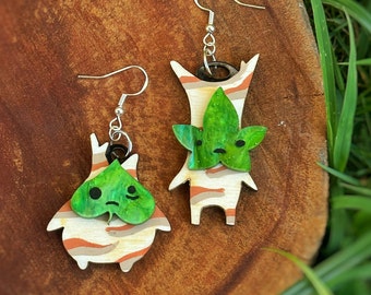 May include: A pair of wooden earrings featuring a green and white striped design with a green leaf on top. The earrings are shaped like a small character with a sad face.