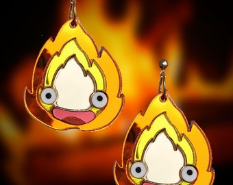 May include: A pair of gold-colored earrings shaped like fire with a cartoon face inside. The earrings are made of acrylic and have a shiny finish.