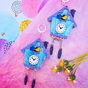 May include: A pair of blue and purple cuckoo clock earrings with a yellow bird and a white clock face. The earrings have a silver chain with a dangling purple and silver glitter charm.