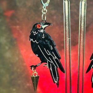 May include: A pair of black raven earrings with red eyes and a gold dangling triangle charm. The earrings are hanging from a silver hook.