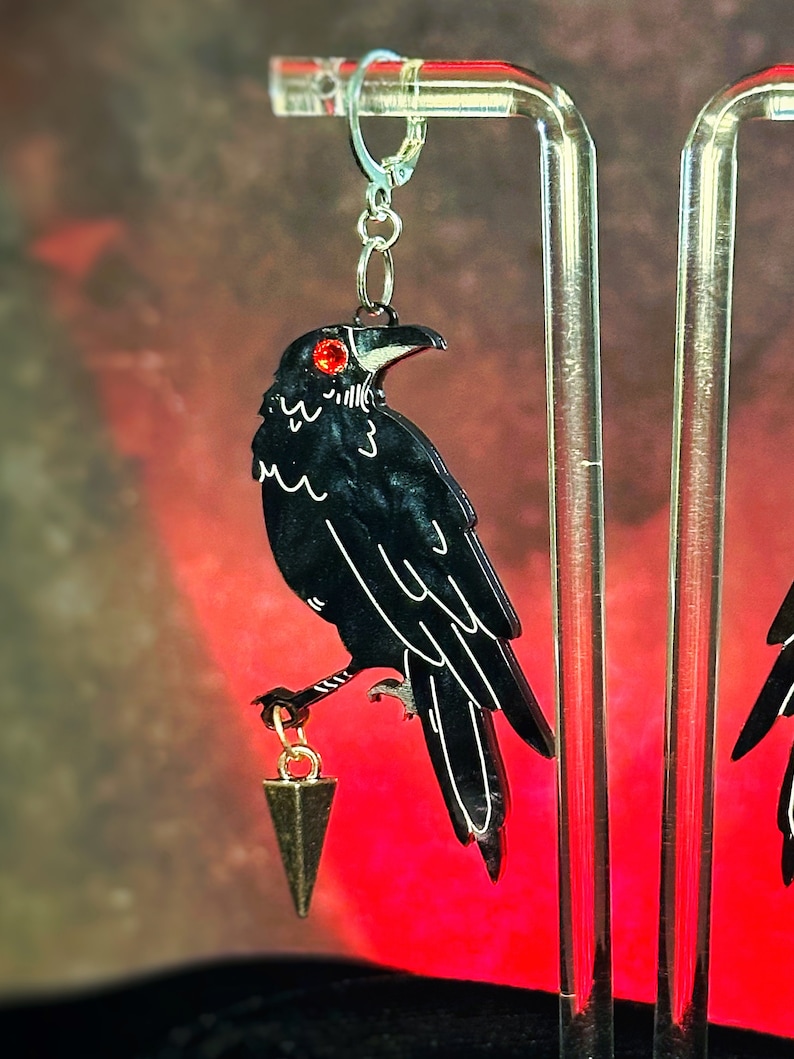 May include: A pair of black raven earrings with red eyes and a gold dangling triangle charm. The earrings are hanging from a silver hook.