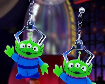 May include: A pair of silver earrings featuring green three-eyed alien characters from the movie Toy Story. The aliens are wearing blue and white striped jumpsuits and are waving.