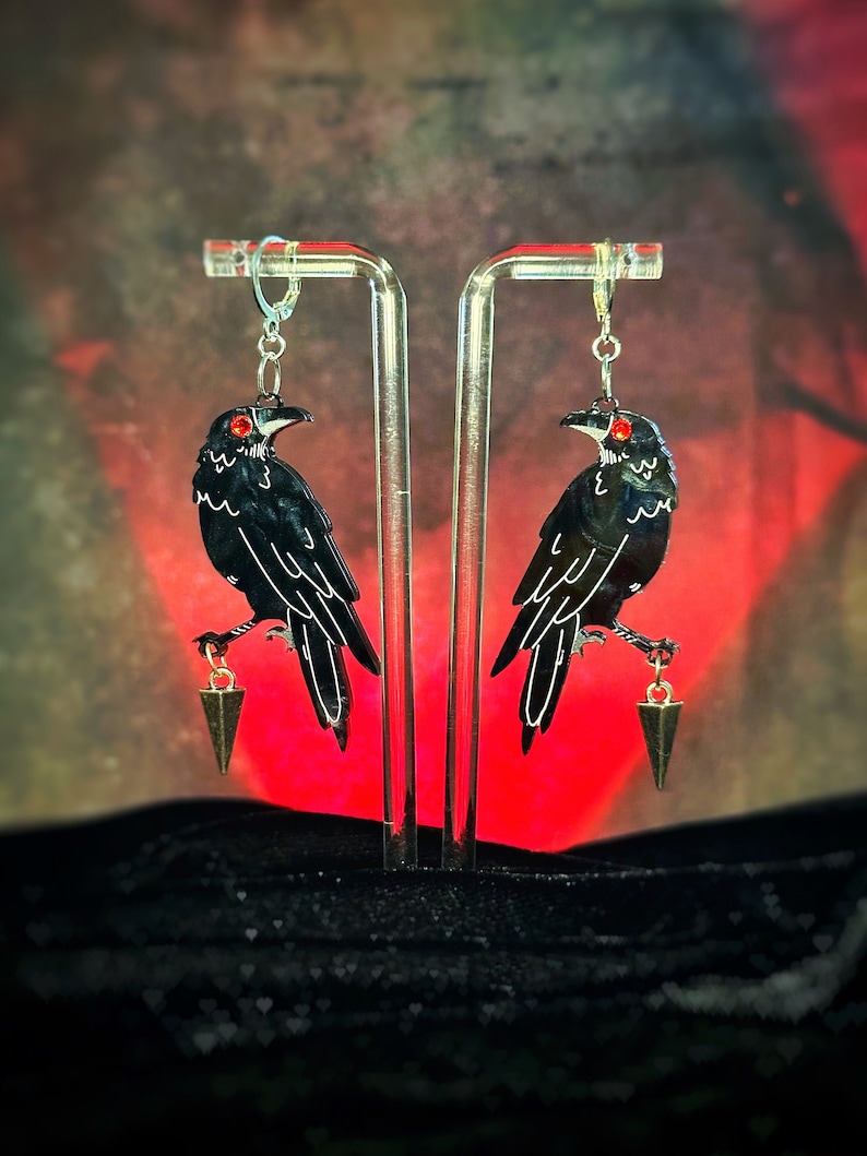May include: A pair of black raven earrings with red eyes and gold dangling charms. The earrings are displayed on a clear acrylic stand against a red and black background.