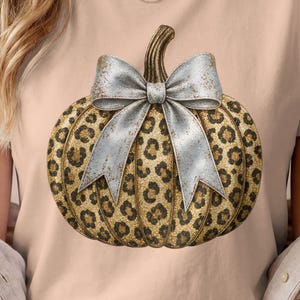 Könnte beinhalten: Ein hellpfirsichfarbenes Kurzarm-T-Shirt mit einer zentralen Grafik eines Kürbisses mit gold-schwarzem Leopardenmuster. Der Kürbis ist mit einer großen, strukturierten silberfarbenen Schleife verziert, die am oberen Stiel gebunden ist.