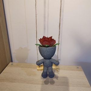 May include: A gray ceramic planter shaped like a person sitting on a swing. The planter has a red flower inside. The swing is made of twine.