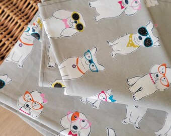 Könnte beinhalten: Graue Stoffservietten mit einem sich wiederholenden Muster aus weißen Cartoon-Hunden, die Sonnenbrillen und Brillen in verschiedenen Farben tragen. Die Hunde haben verschiedenfarbige Halsbänder und Fliegen. Die Servietten sind gefaltet und in einem Weidenkorb gestapelt.