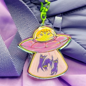 Könnte beinhalten: Ein farbenfroher Acryl-Anhänger mit einem Cartoon-UFO mit einem Katzenpiloten und drei lila Katzen darunter. Das UFO ist rosa mit gelben und grünen Konturen, und die Katzen haben gelbe Augen. Der Anhänger ist an einer grünen Kette befestigt.