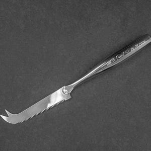 Könnte beinhalten: Ein silberfarbenes Käsemesser aus Edelstahl mit gezahnter Klinge und zweizinkiger Spitze. Der Griff ist mit dem Text "Lea & Daniel", einem Herzsymbol und dem Datum "26.09.2026" in einer kursiven Schriftart graviert.