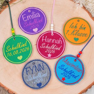 Könnte beinhalten: Sechs kreisförmige Filzanhänger in Lila, Grün, Pink, Orange, Grau und Blau, jeweils bestickt mit Namen wie Emilia, Hannah, Fynn und Maximilian sowie dem Text Schulkind 2026 oder Ich bin 1. Klasse und kleinen Herz- oder Sternsymbolen.