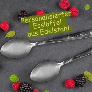 Könnte beinhalten: Zwei personalisierte Esslöffel aus Edelstahl mit graviertem Text. Die Löffel liegen auf einer dunkelgrauen Oberfläche mit roten und schwarzen Beeren und grünen Blättern.