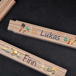 Könnte beinhalten: Zwei Holz-Klapplineale mit unterschiedlichen Designs. Ein Lineal zeigt ein Bauthema mit gelben LKWs und dem Namen "Finn" darauf gedruckt. Das andere Lineal zeigt ein Werkzeugthema mit einem Pinsel, einem Hammer und anderen Werkzeugen und dem Namen "Lukas" darauf gedruckt.