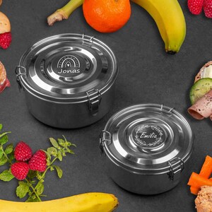 Könnte beinhalten: Zwei Edelstahl-Lunchboxen mit personalisierten Namen, umgeben von Lebensmitteln wie Sandwiches, Obst und Keksen. Die Behälter haben ein kreisförmiges Design mit einem sicheren Verschluss. Der Name "Jonas" ist sichtbar.