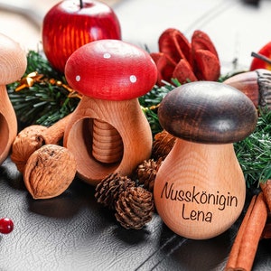 Könnte beinhalten: Drei Nussknacker aus Holz in Form von Pilzen, einer rot, einer braun und einer aus Naturholz. Die Nussknacker sind mit weißen Punkten und dem Text "Nusskönigin Lena" verziert. Die Nussknacker sind von Tannenzweigen, Zimtstangen und Tannenzapfen umgeben.