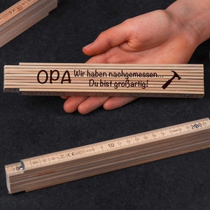 Könnte beinhalten: Ein Holz-Klappmaßstab mit der Aufschrift "OPA Wir haben nachgemessen... Du bist großartig!" und einem kleinen Hammer-Symbol. Der Maßstab ist geschlossen und wird in einer Hand gehalten.