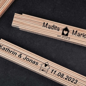 Könnte beinhalten: Drei Holz-Klapplineale mit personalisierten Gravuren. Das erste Lineal lautet "Madita & Mario est. 2023" mit einem Haussymbol und einem Herzen. Das zweite Lineal lautet "Kathrin & Jonas 11.08.2023" mit einem Haussymbol und einem Herzen. Das dritte Lineal ist leer.