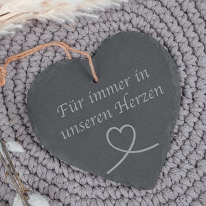 Könnte beinhalten: Eine schwarze, herzförmige Schiefertafel mit dem Text "Für immer in unseren Herzen" und einem kleinen Herzsymbol, das darauf graviert ist. Die Tafel hängt an einer grauen gehäkelten Oberfläche.
