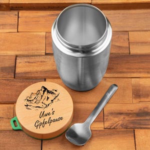 Könnte beinhalten: Ein Edelstahl-Essensbehälter mit Holzdeckel und kleinem Löffel. Der Deckel zeigt eine Berg-Grafik und den Text "Uwe's Gipfelpause". Der Behälter ist offen und zeigt sein Inneres.