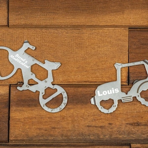 Könnte beinhalten: Zwei silberne Multi-Tool-Fahrrad-Gadgets mit den Texten "Bernd's Multitalent" und "Louis". Die Werkzeuge liegen auf einer Holzoberfläche.