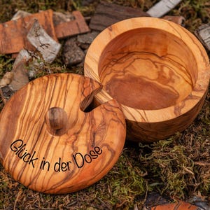 Könnte beinhalten: Eine Holzkiste mit Deckel aus Olivenholz. Der Deckel hat einen kleinen Knopf und die Inschrift "Glück in der Dose" ist in den Deckel graviert.