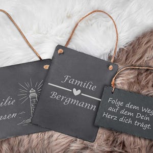 Könnte beinhalten: Drei schwarze Schiefertafeln mit weißem Text und Bildern hängen an einer Schnur. Die Schilder zeigen "Familie Hansen" mit einem Leuchtturm, "Familie Bergmann" mit einem Herzen und "Folge dem Weg auf dem Dein Herz dich trägt." mit einem Löwenzahn.
