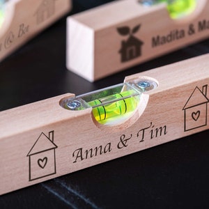 Könnte beinhalten: Ein Holz-Wasserwaage mit einer grünen Blase und einem Haus mit einem Herz-Design, das darauf graviert ist. Die Wasserwaage ist mit dem Text "Anna & Tim" personalisiert.
