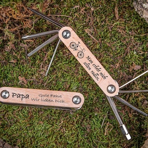 Könnte beinhalten: Ein Holz-Multitool mit einem Fahrrad-Design und dem Text "Man erlebt mich selten radlos." Das Werkzeug hat einen Holzgriff und eine Metallklinge. Auf der anderen Seite des Werkzeugs steht der Text "Papa Gute Fahrt Wir lieben Dich..."