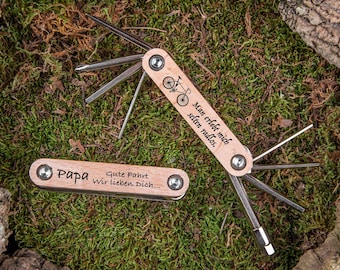 May include: A wooden multi-tool with a bicycle design and the text "Man erlebt mich selten radlos."  The tool has a wooden handle and a metal blade.  The other side of the tool has the text "Papa Gute Fahrt Wir lieben Dich..."